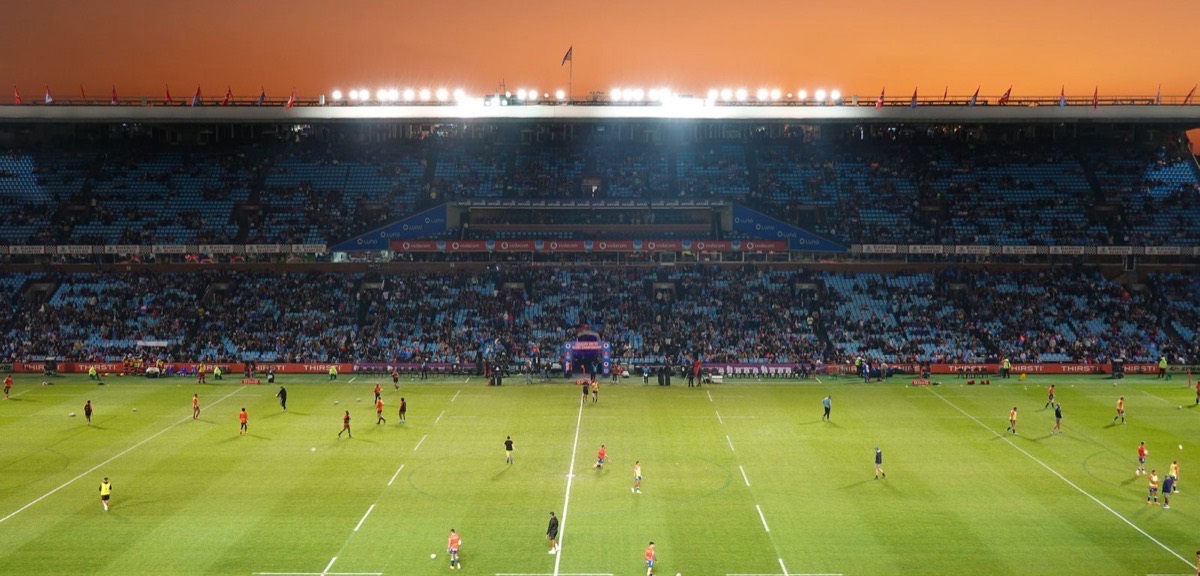 A floodlit stadium at sunset with players warming up on the pitch