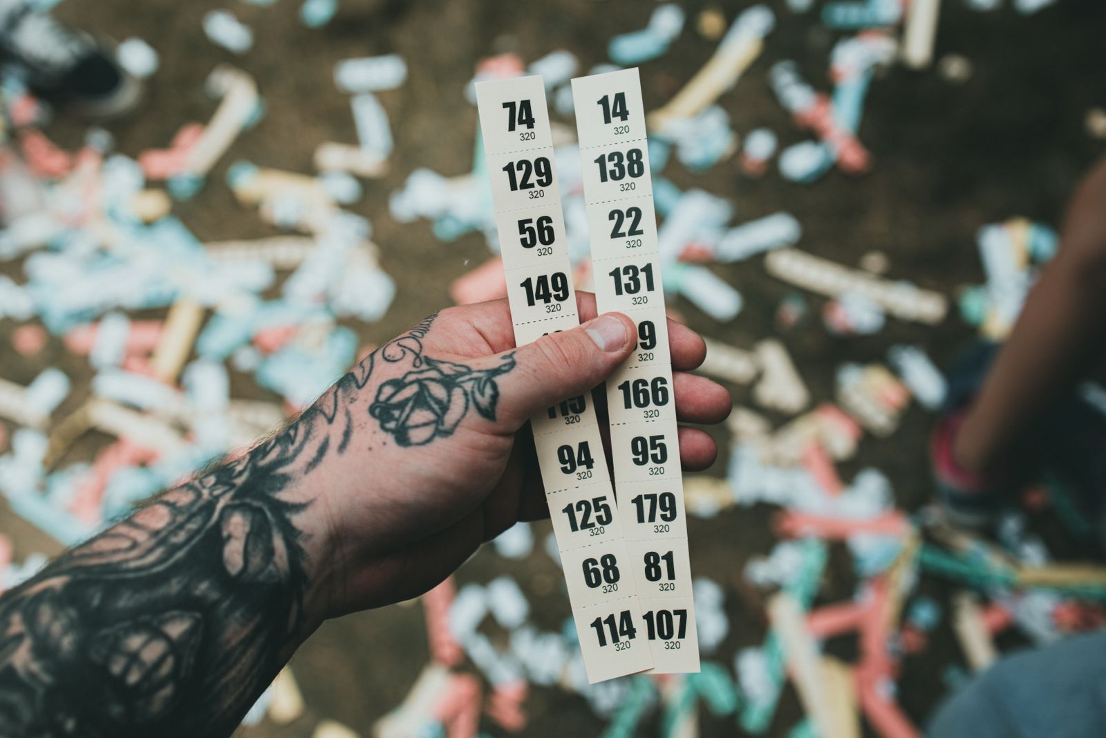 A tattooed hand holding up two strips of numbered raffle tickets over a ground scattered with confetti at an outdoor event