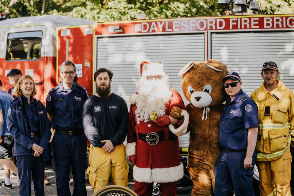 Daylesford CFA volunteers lined up with Santa and a teddy bear mascot in front of a fire truck at the Cliffy's Emporium Christmas raffle